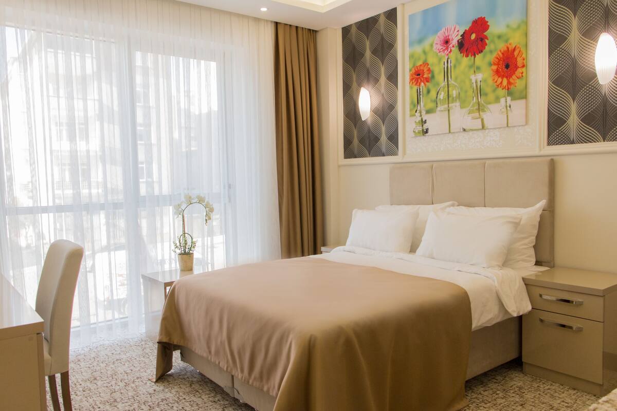 Habitación de hotel con cama, mesita de noche, silla y un ventanal con cortinas translúcidas.