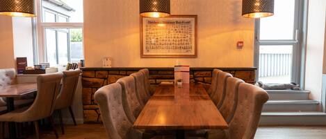 Un restaurant avec une longue table en bois, des chaises rembourrées et des lampes suspendues.