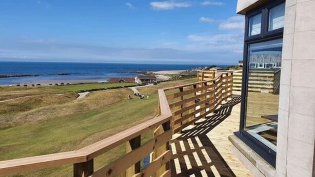 A balcony with a wooden railing overlooking a grassy area and the sea. - Golf View Hotel (Lossiemouth)