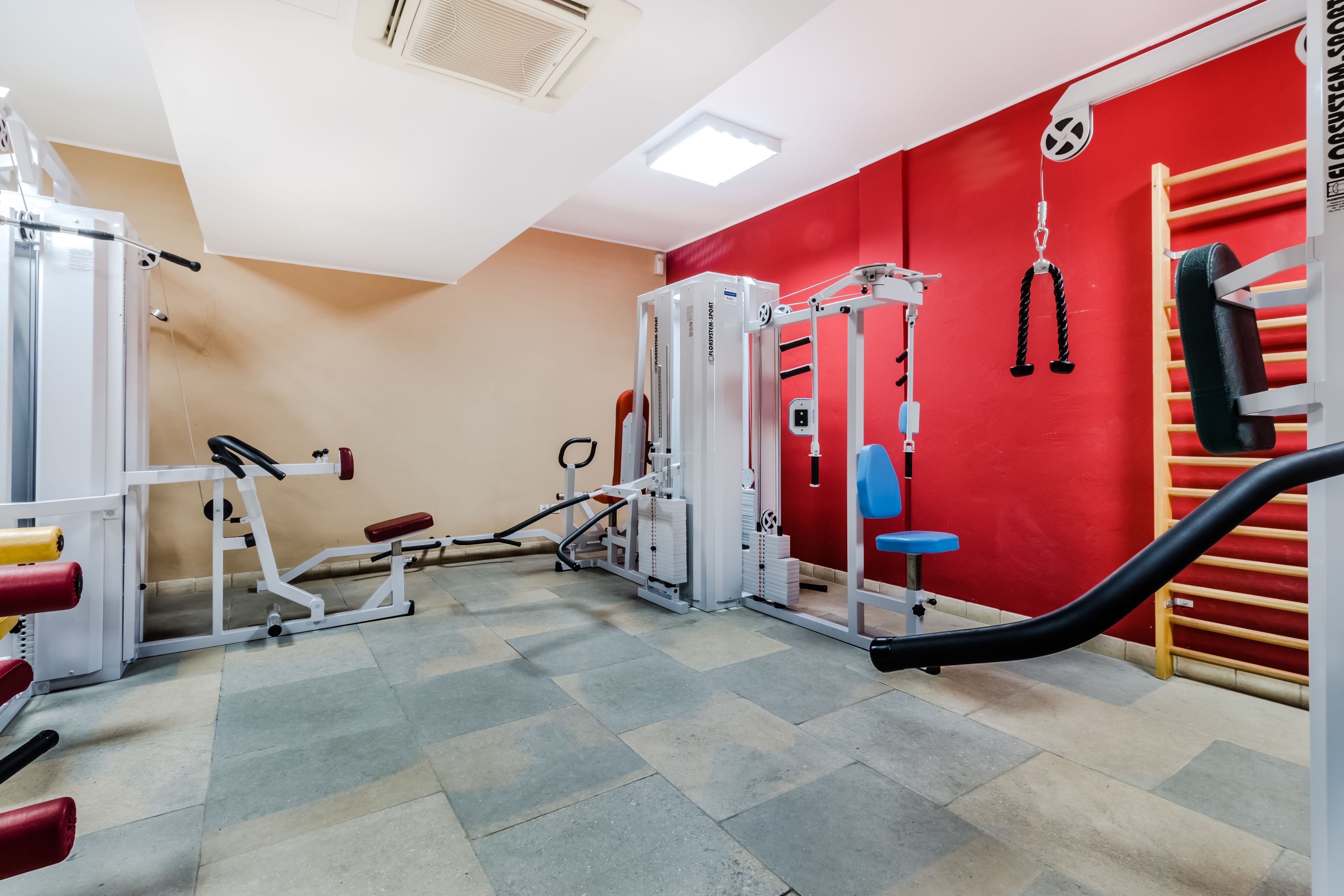 Une salle de sport équipée de divers appareils d’exercice, avec un mur rouge et un ventilateur au plafond.