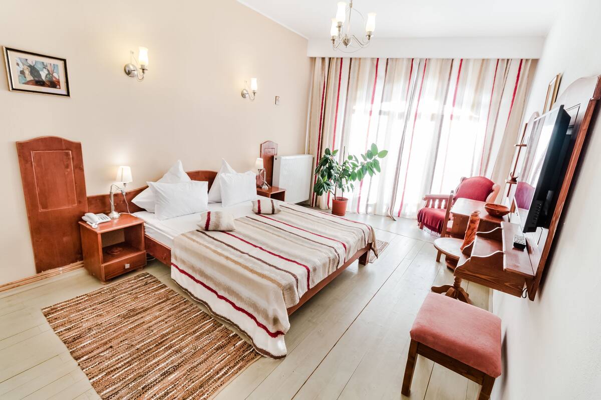Ein Hotelzimmer mit einem großen Bett, Nachttischen, einem Schreibtisch, einem Sessel, einem Spiegel und einer Zimmerpflanze.