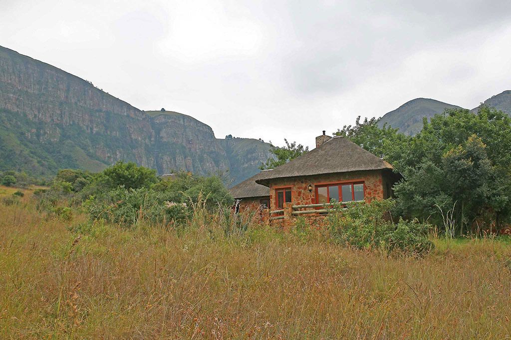 A rustic stone building with a thatched roof, surrounded by tall grass and trees, set against a backdrop of mountains.