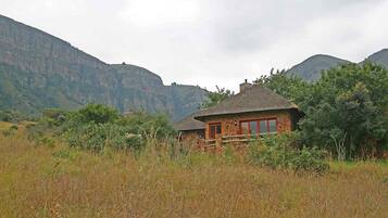 A rustic stone building with a thatched roof, surrounded by tall grass and trees, set against a backdrop of mountains.