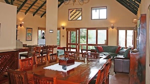 A spacious dining area with a long wooden table, surrounded by wooden chairs. There is a chandelier hanging from the ceiling, and large windows allowing natural light to enter.