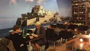 Een restaurant met een muurschildering van een kuststad, houten plafond en een wijnrek.