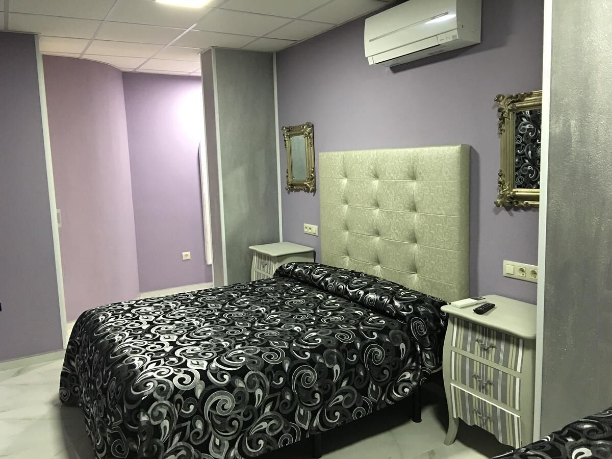 A bedroom with a tufted headboard, a bed with a patterned bedspread, a bedside table with a remote control, and a small cabinet.