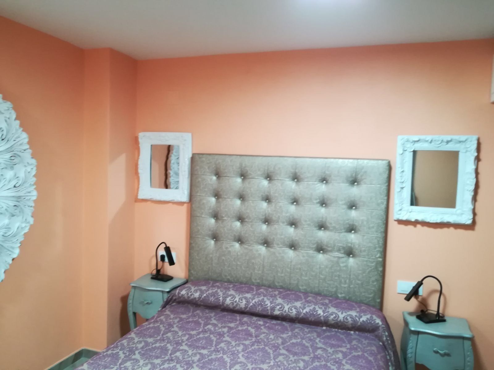 A bedroom with a tufted headboard, a purple patterned bedspread, two bedside tables with lamps, and decorative mirrors on the walls.