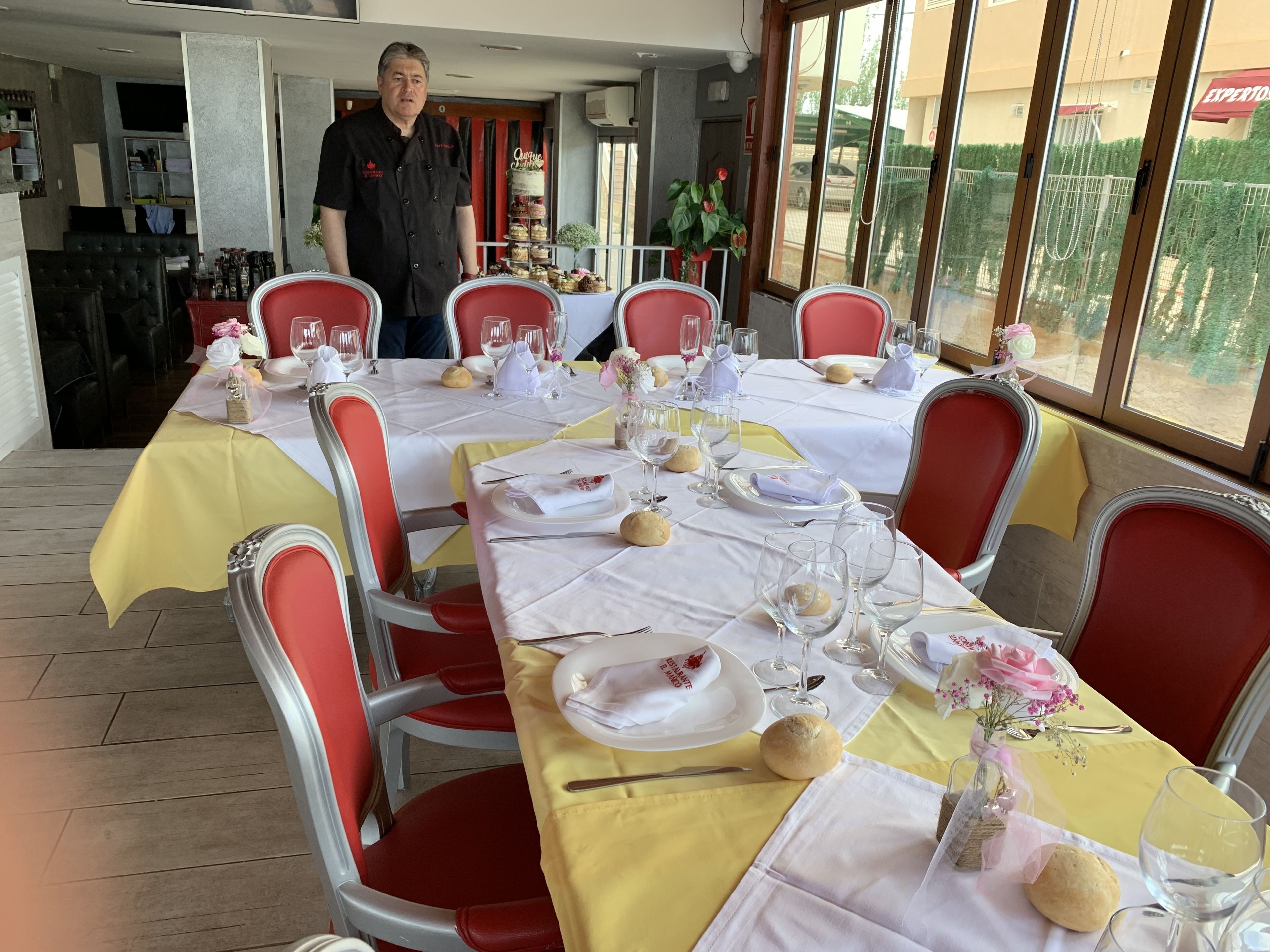 Un restaurante con una larga mesa dispuesta para una comida, sillas rojas y un camarero cerca.
