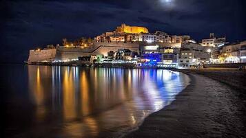 Une ville côtière de nuit, avec une imposante structure en forme de château qui domine une colline, une plage paisible au premier plan et des bâtiments illuminés le long de la côte.