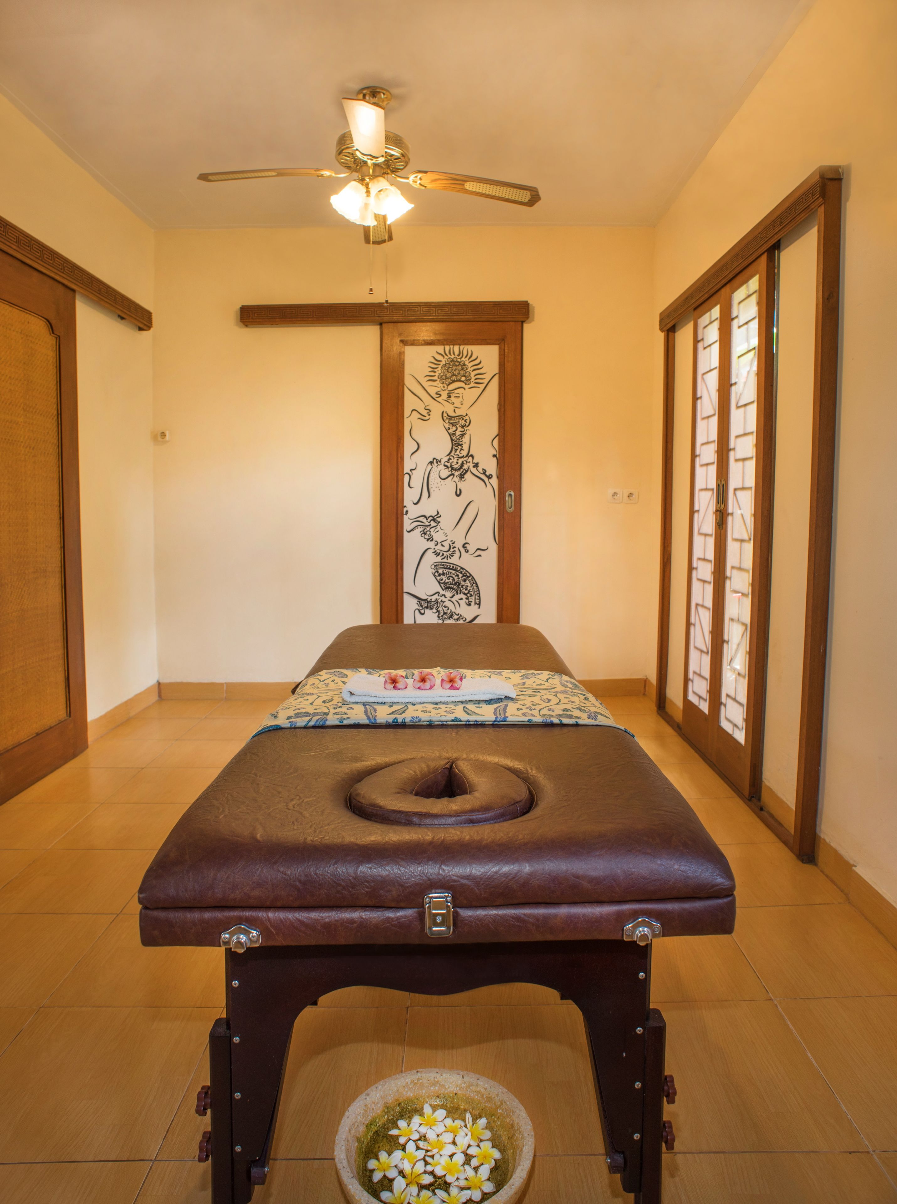 A massage table with a headrest, a small bowl of flowers, and a ceiling fan in a room with a wooden door and a tiled floor.