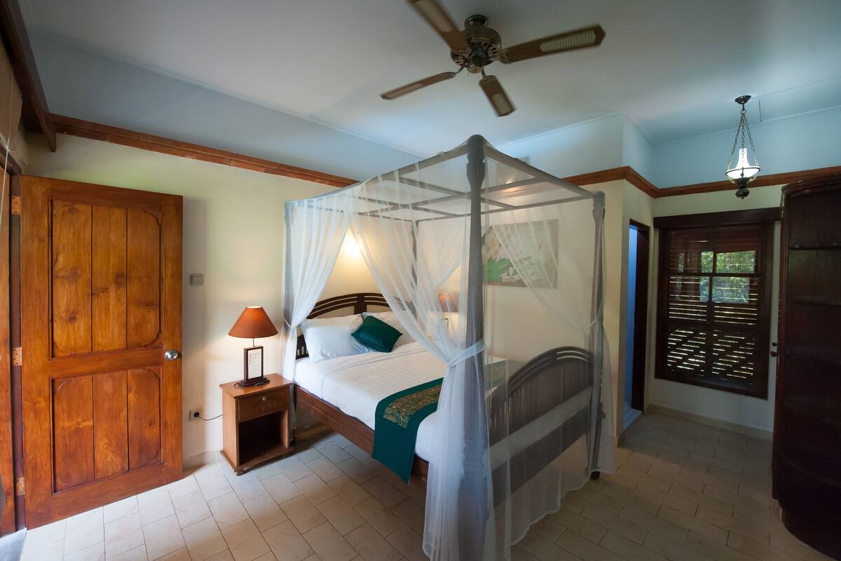 Een kamer met een hemelbed, een plafondventilator, een nachtkastje met lamp en een houten deur.