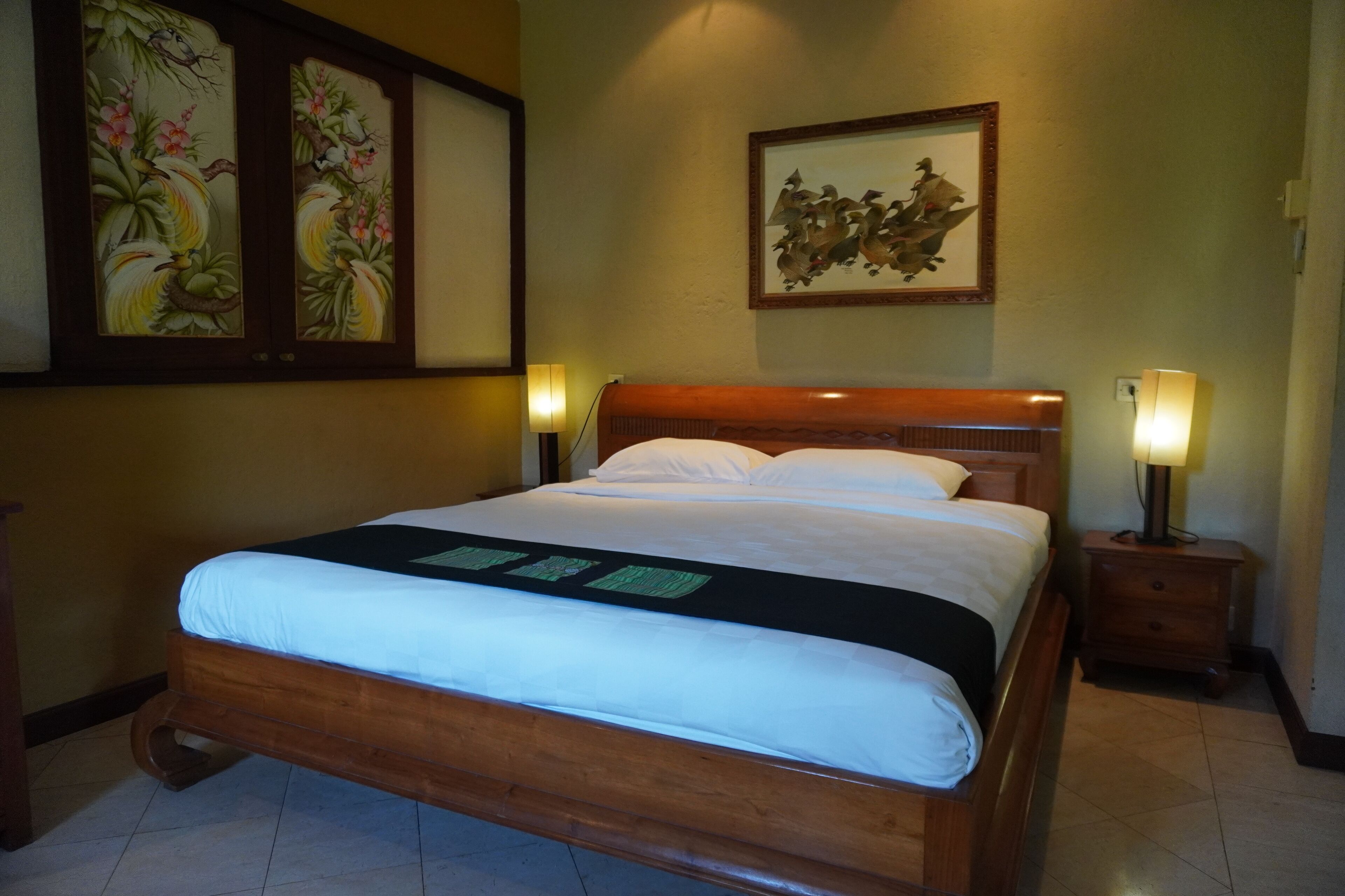 Una cama bien tendida con un cabecero de madera, dos mesitas de noche con lámparas, y obras de arte en las paredes.