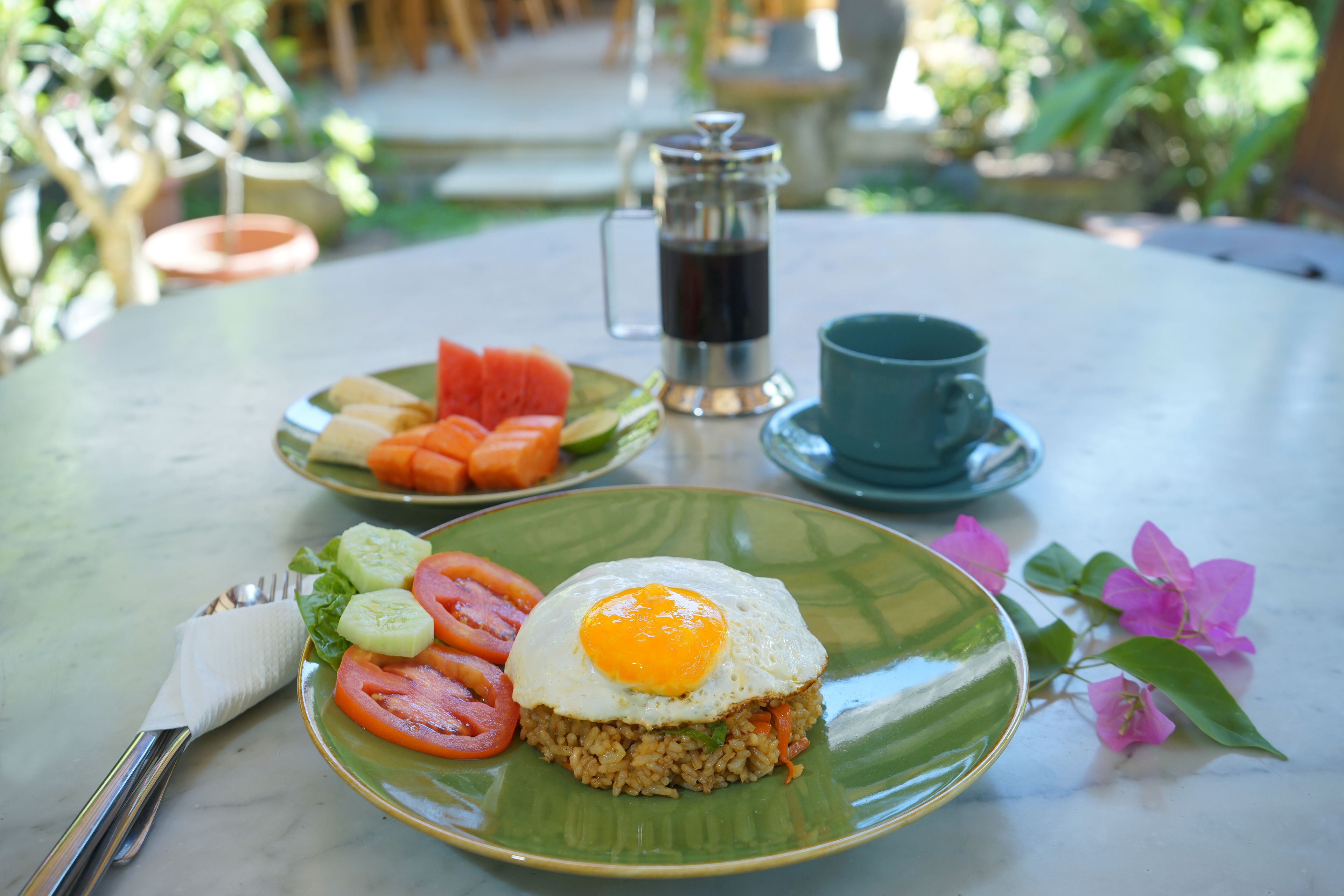Una mesa con arroz frito coronado por un huevo frito, una taza de café y un plato de vegetales en rodajas.