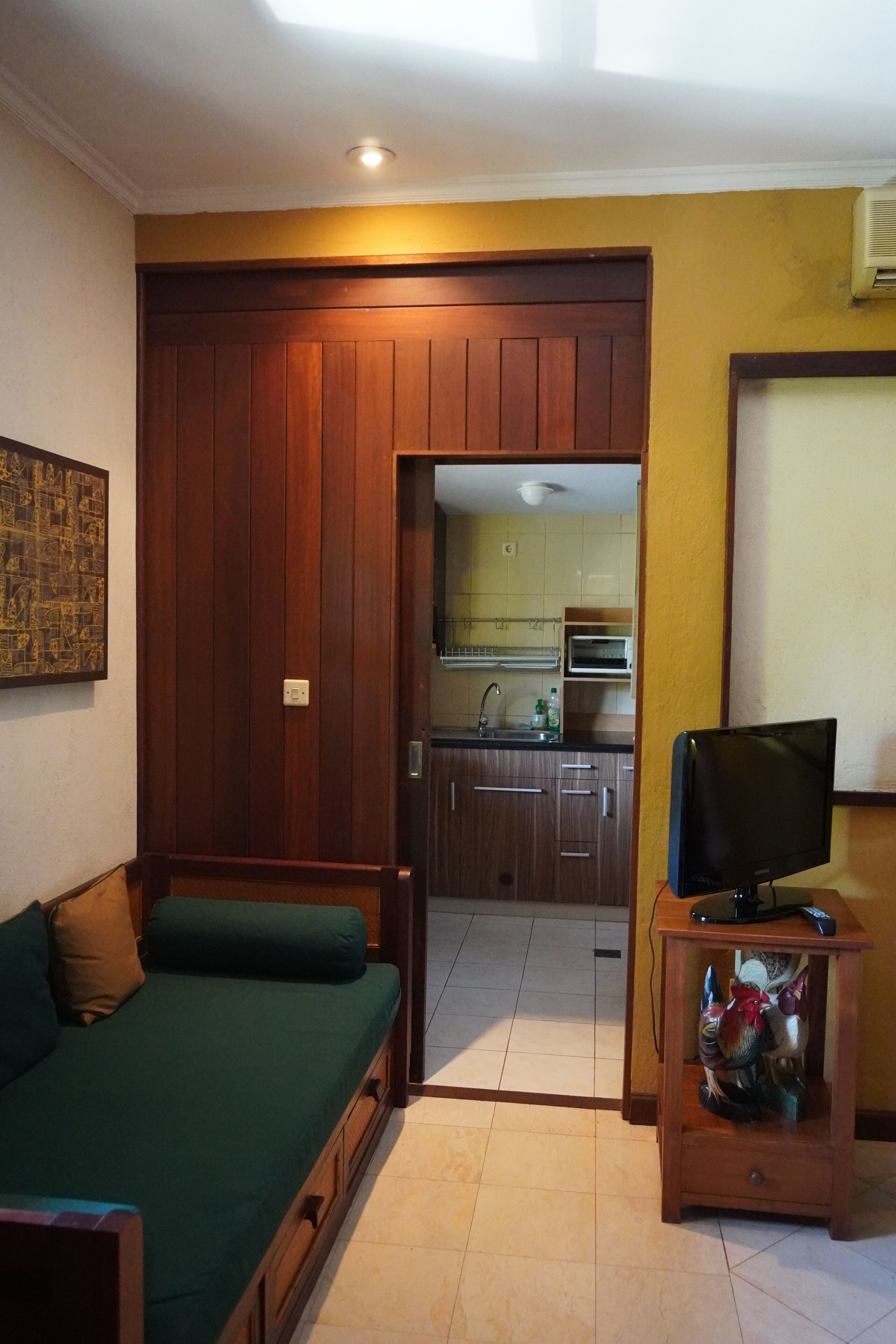 A compact living space with a sofa, a television on a wooden stand, a kitchen area visible through an open door, and a ceiling light.