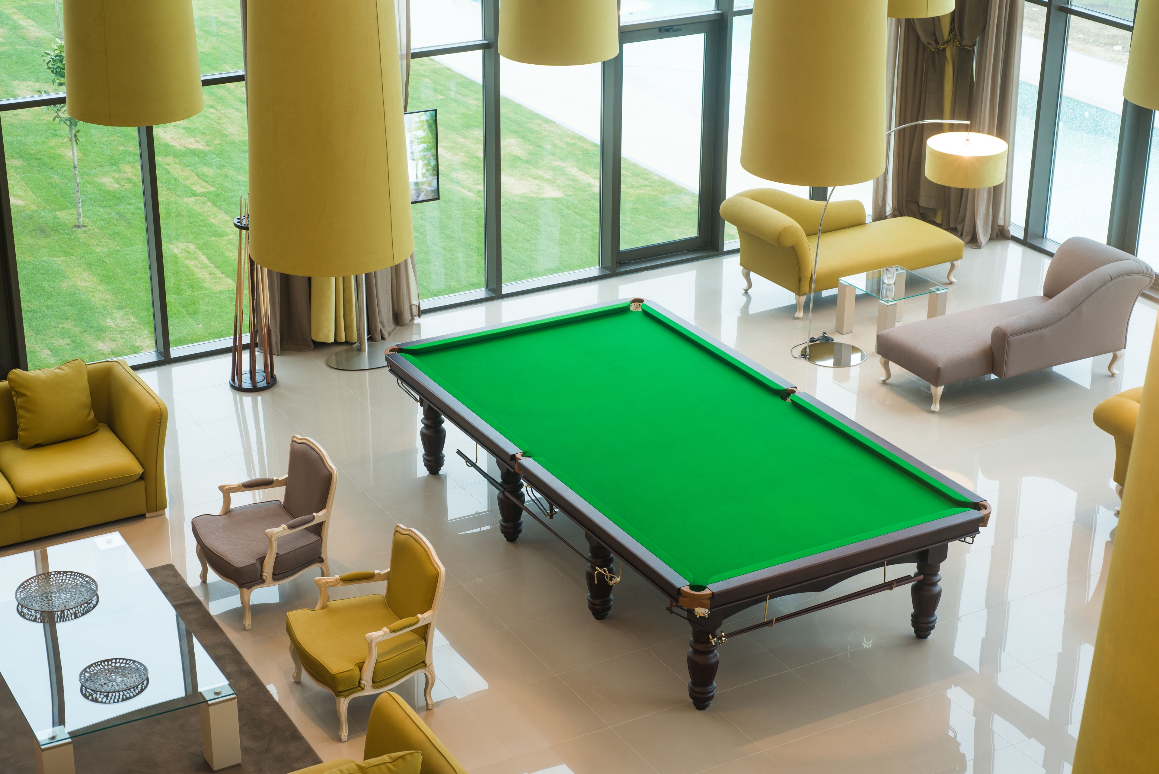 A pool table in a well-lit room with large windows, yellow chairs, and a glass coffee table.
