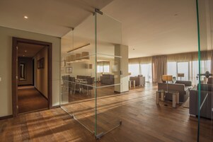 Una habitación de hotel moderna con separador de vidrio, suelo de madera, zona de comedor con sillas y mesa, y una sala de estar con sofás y una lámpara.