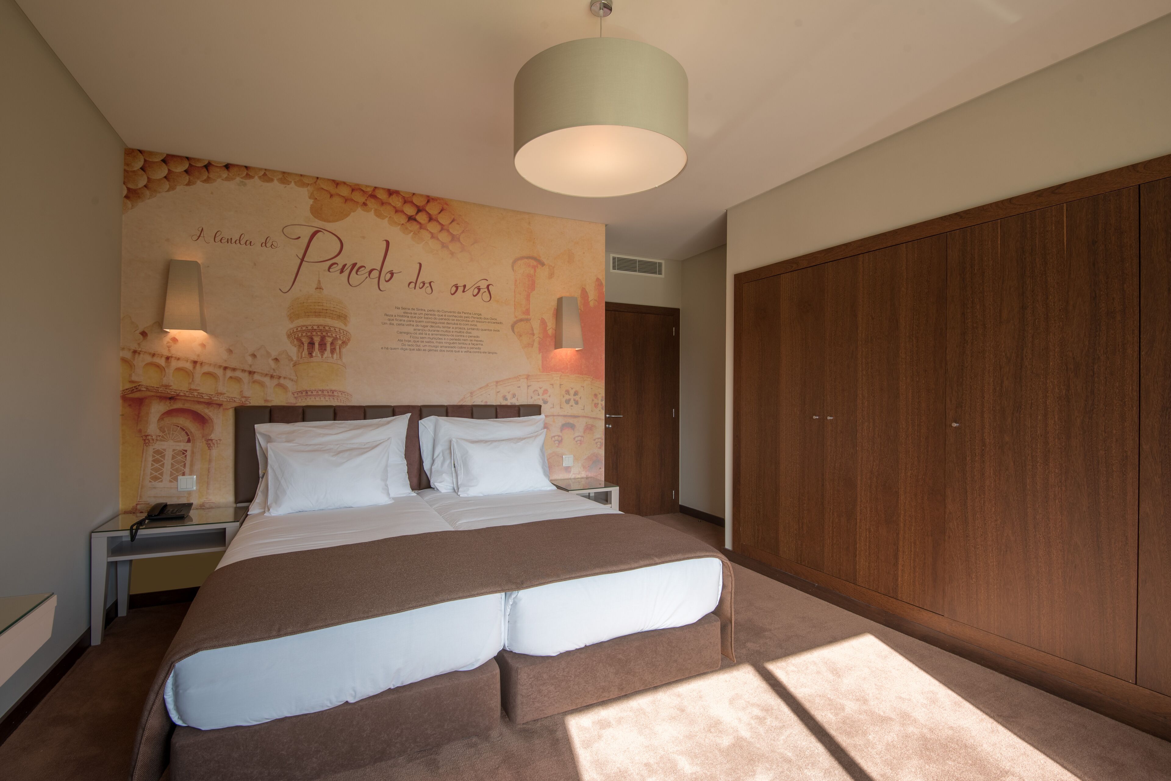 A hotel room with a large bed, a wooden wardrobe, a wall with a decorative mural, and a hanging light fixture.
