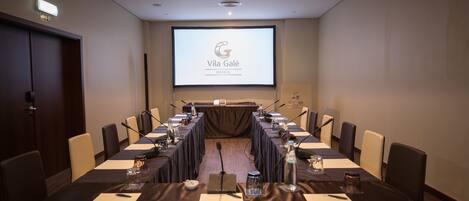 A conference room with a long table, chairs, a projector screen displaying 'Vila Galé Hotelis', and a podium.