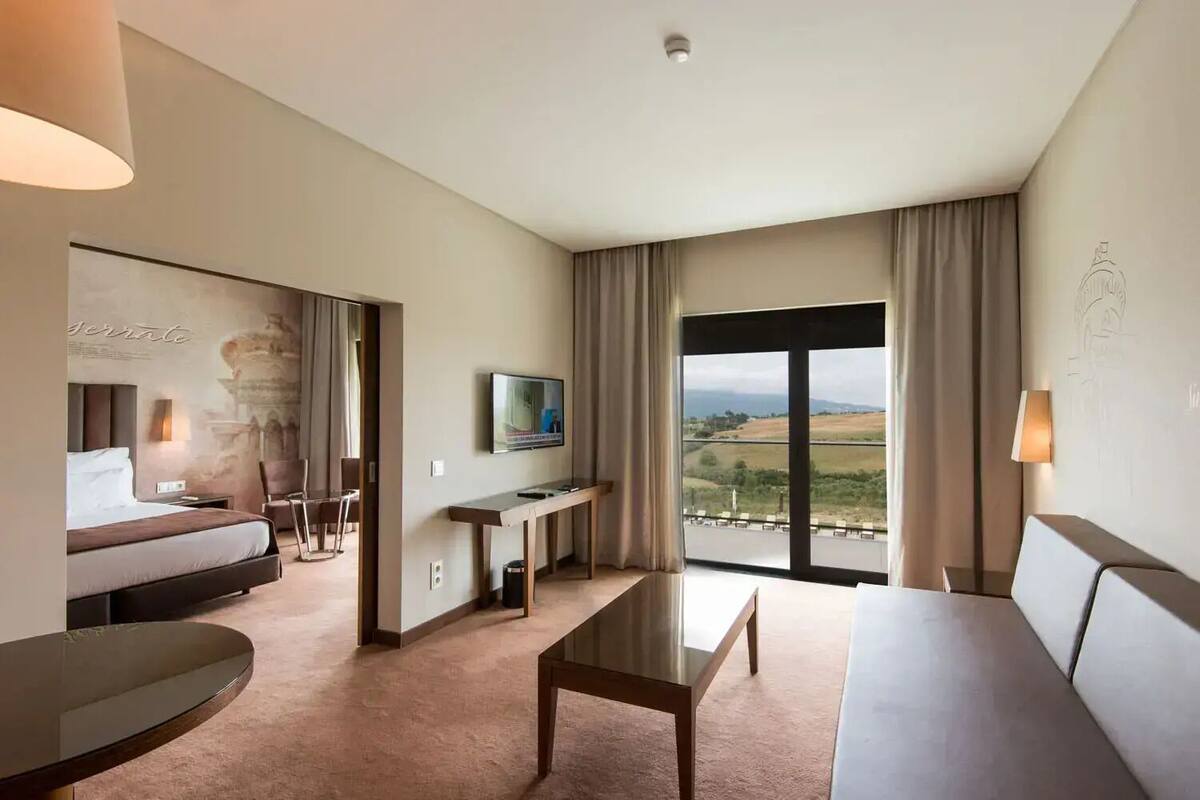 Ein Hotelzimmer mit Bett, Sofa, Esstisch, Fernseher und einem großen Fenster mit Blick auf die Landschaft.