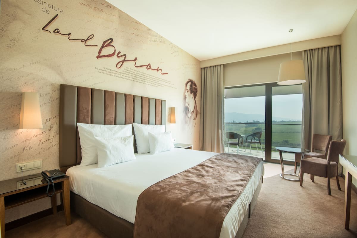 Ein Hotelzimmer mit einem großen Bett, einem Schreibtisch mit Telefon, einem Sessel, einem Tisch und Blick ins Freie.