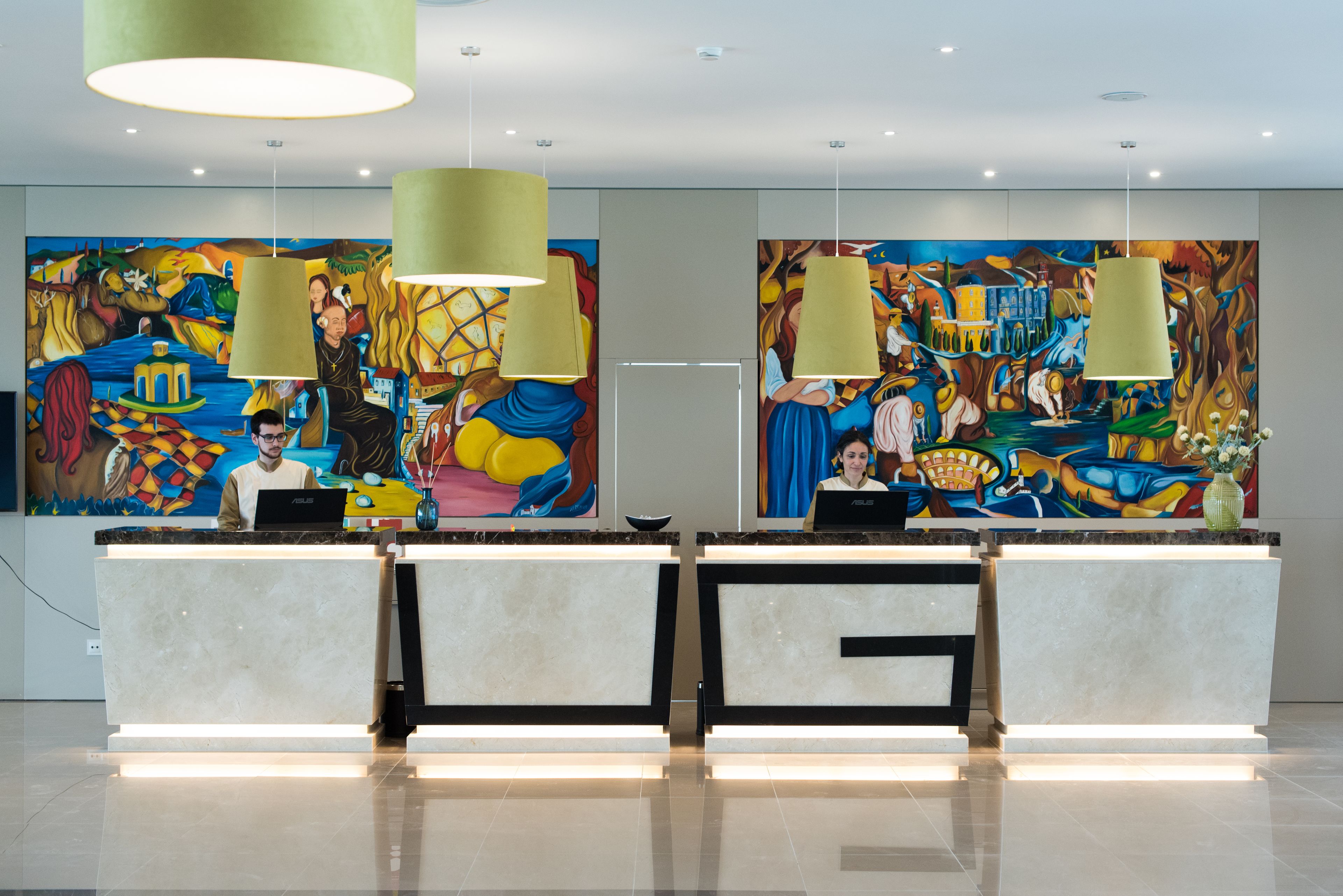 A hotel reception area with two reception desks, each with a laptop, a vase, and a painting on the wall.