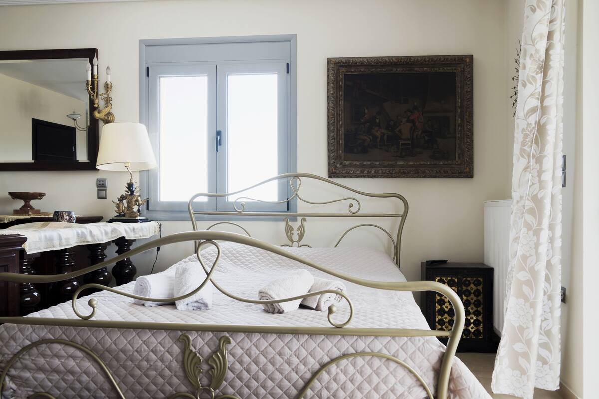 A neatly made bed with a quilted cover, a bedside table with a lamp, a mirror, a framed painting, and a window with curtains.