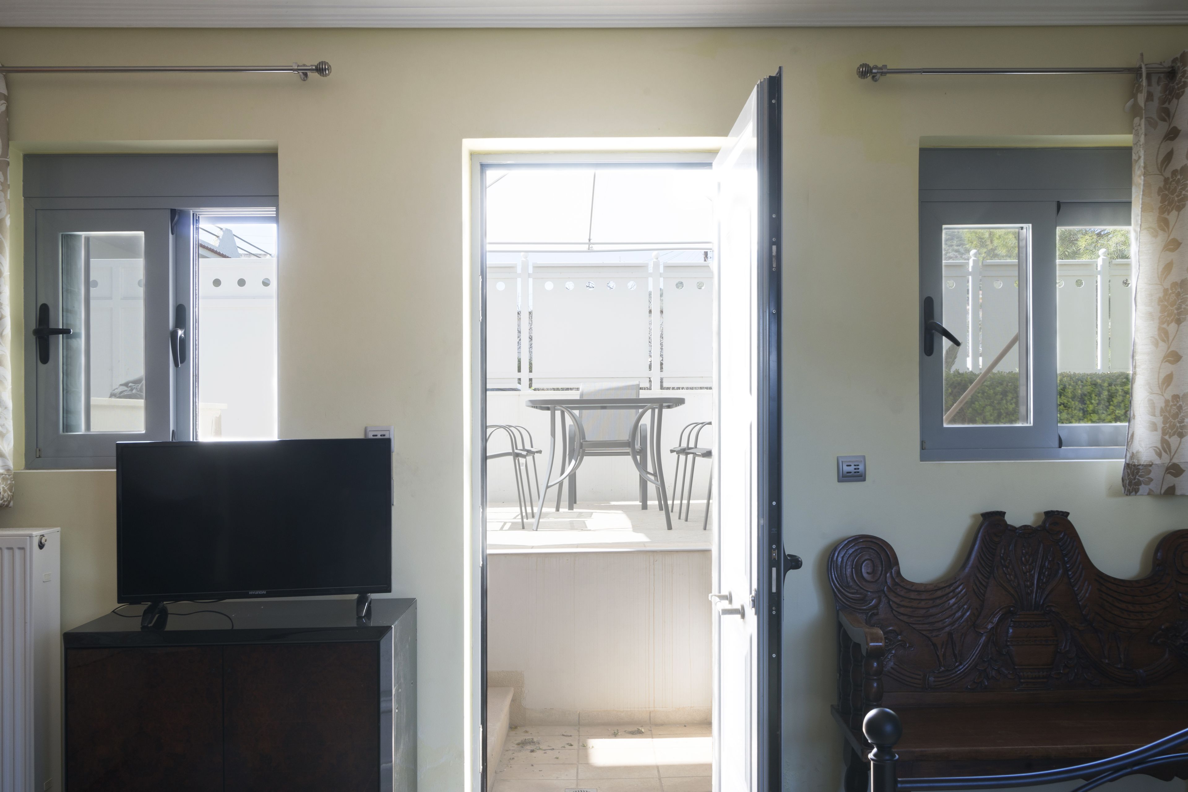 A room with a television on a cabinet, a wooden bed with a headboard, and an open door leading to a balcony with a table and chairs.
