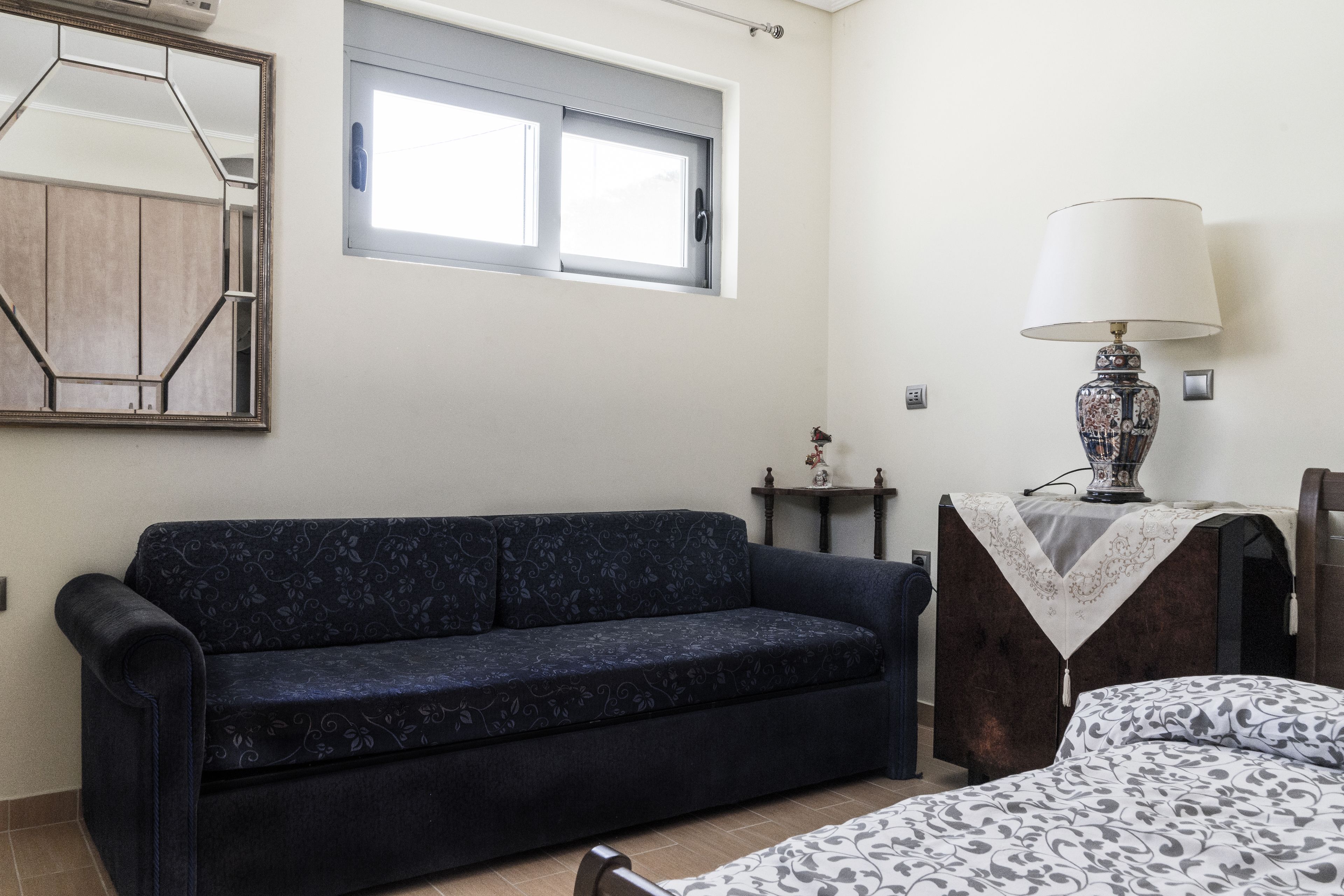 Habitació amb un sofà blau fosc, un llit amb roba de llit estampada, una taula amb una làmpada i una finestra amb vista a l'exterior.