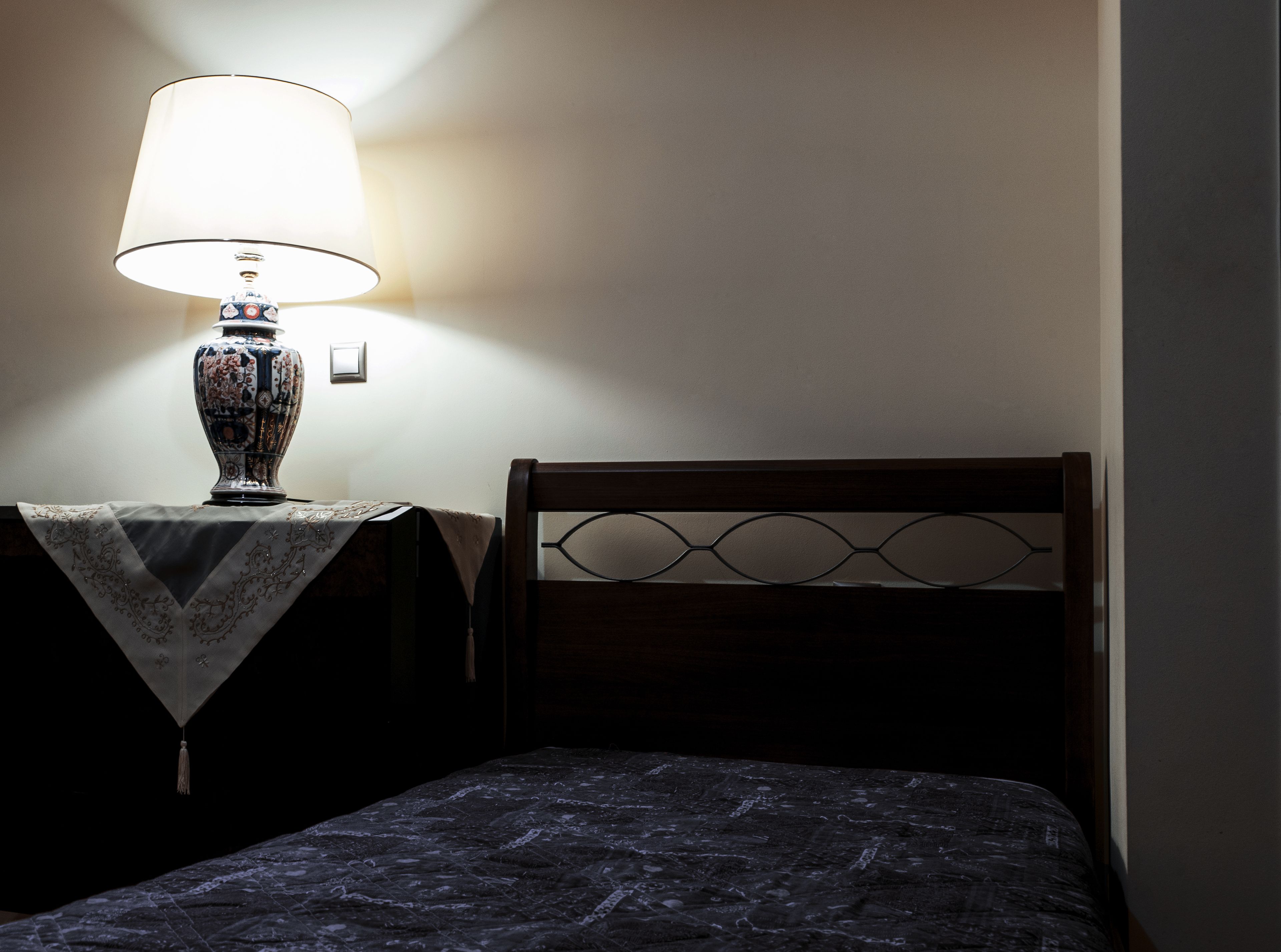 A bed with a dark, textured bedspread, a wooden headboard with a geometric design, and a table with a decorative lamp and a small vase.