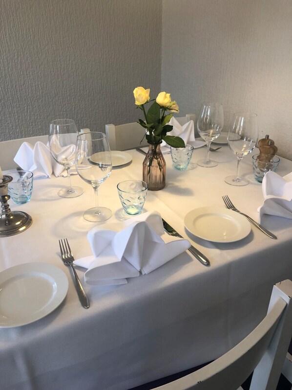 Mesa redonda para comer con mantel blanco, platos, cubiertos y copas de cristal.