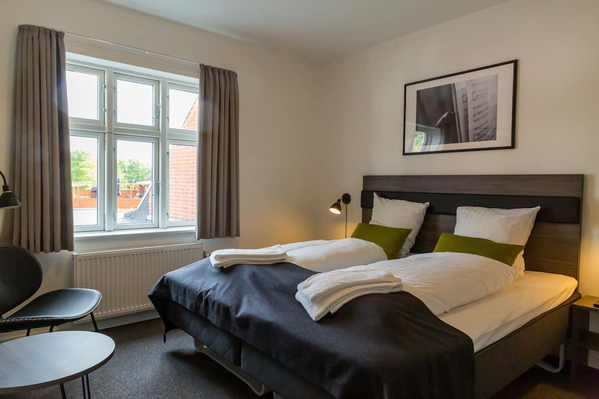 A hotel room with a bed, a chair, a table, a window with curtains, a radiator, and a framed picture on the wall.