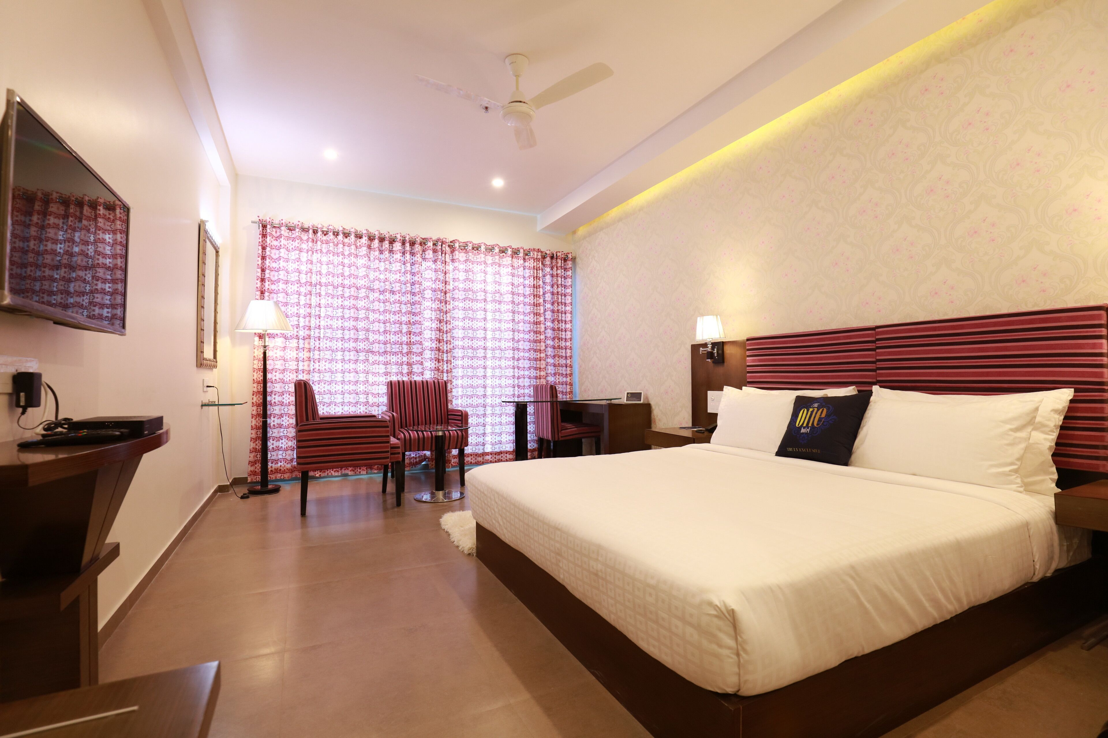 A hotel room with a large bed, a desk with a chair, a television, and a ceiling fan.
