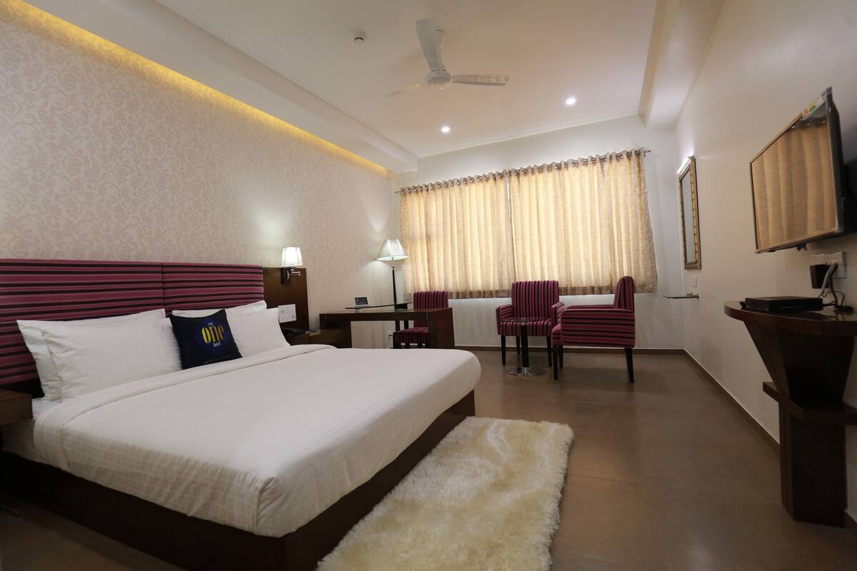 Une chambre d’hôtel avec un grand lit, un bureau avec une lampe, une chaise, une petite table et un téléviseur fixé au mur.