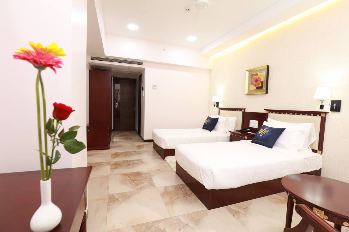 Une chambre d’hôtel avec deux lits simples, une table de chevet en bois, un bureau, un vase avec des fleurs et une porte donnant sur une autre pièce.