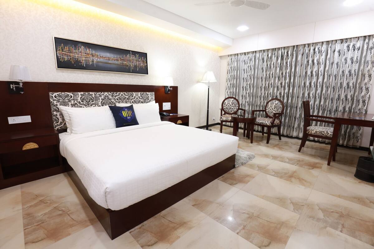 Une chambre d’hôtel avec un grand lit, un bureau avec deux chaises, une lampe sur pied et un tableau représentant un paysage urbain accroché au mur.