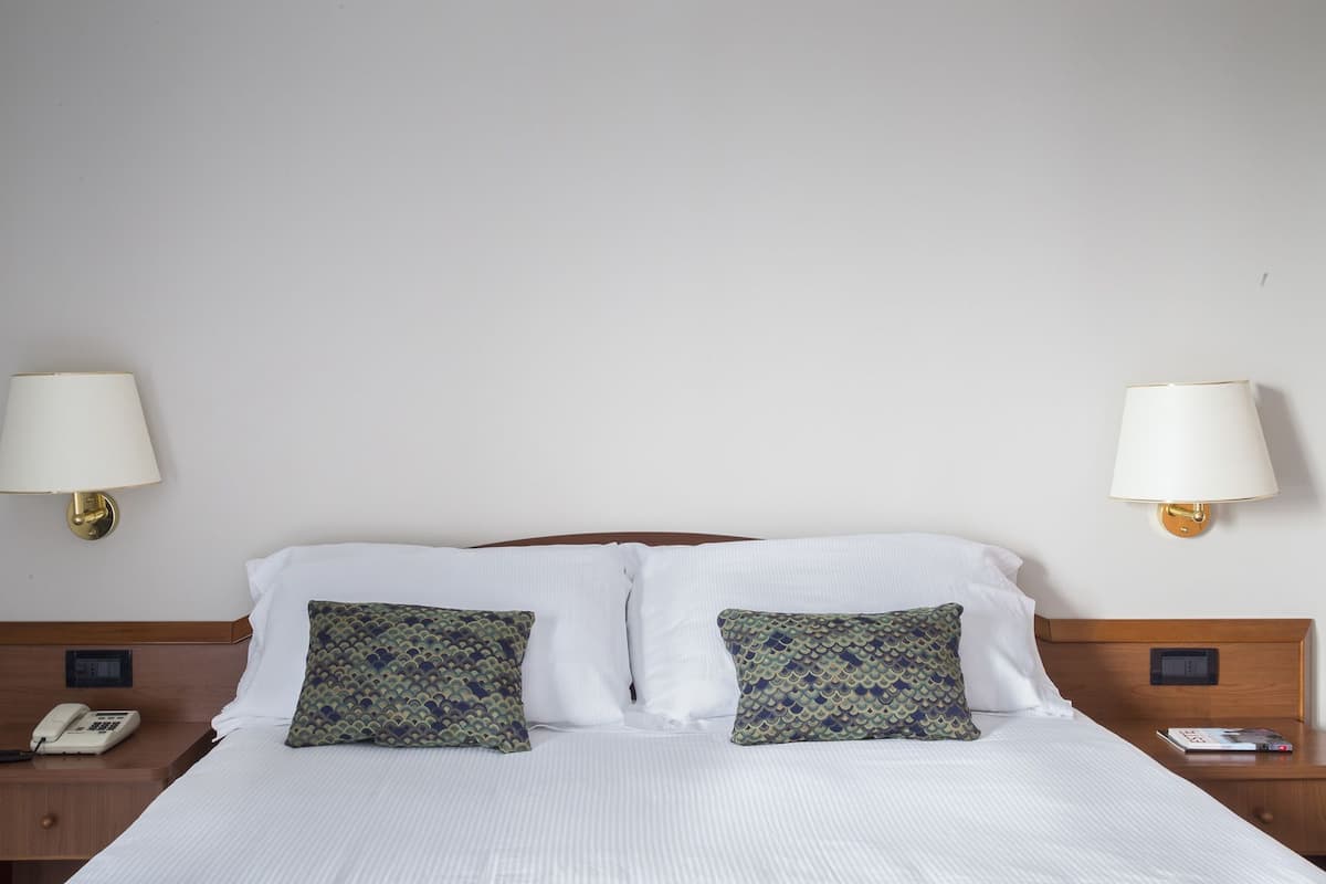 A hotel room with a bed, two pillows, two bedside tables, a telephone, and two wall-mounted lamps.