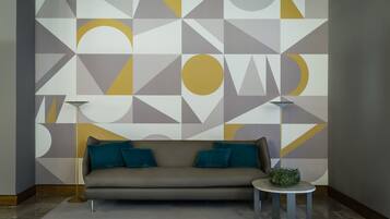 Ein modernes Wohnzimmer mit einer Wand mit geometrischem Muster, einer Couch mit türkisfarbenen Kissen, einem kleinen runden Tisch mit einer Zimmerpflanze und zwei Stehlampen.