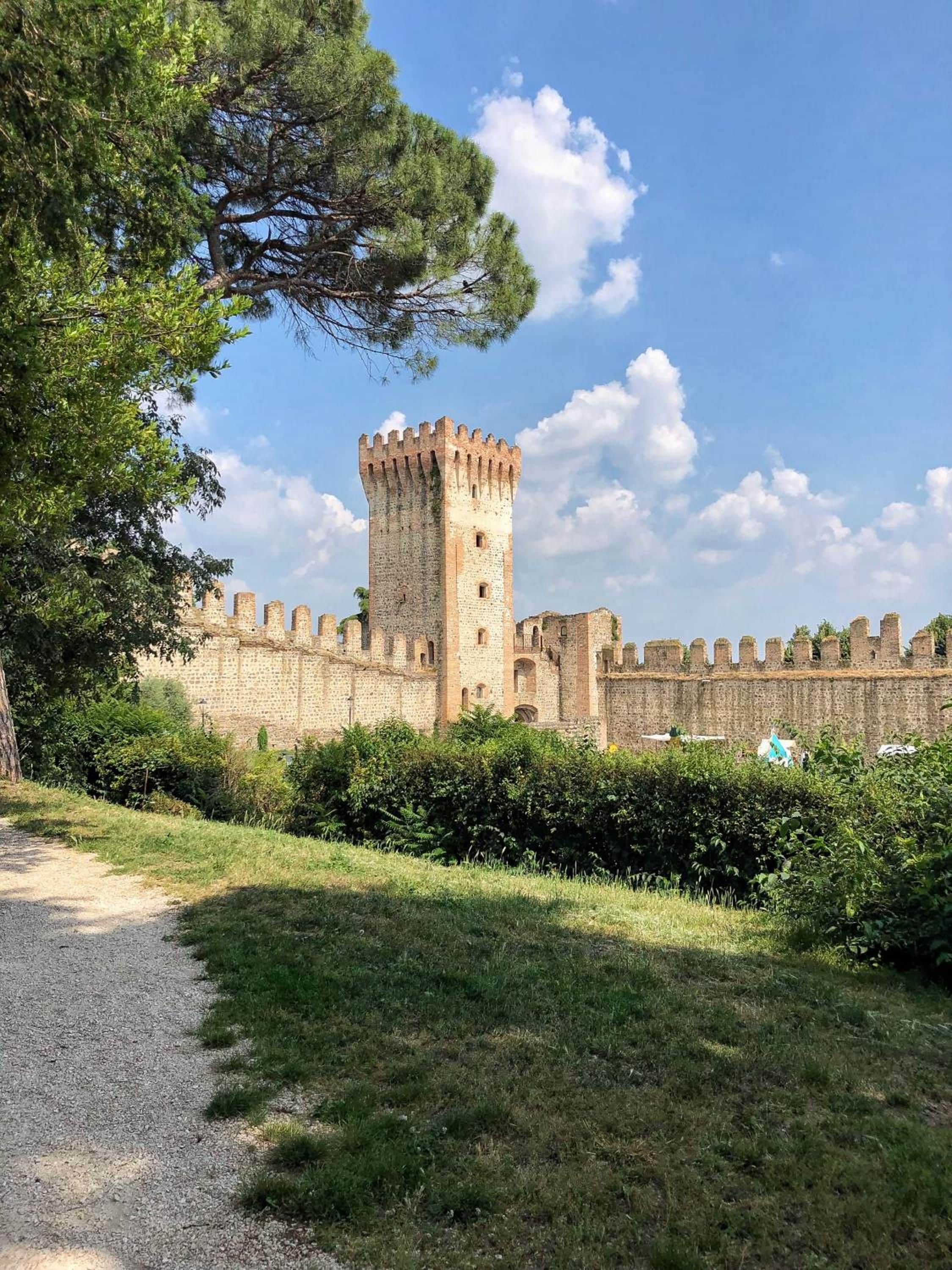 A historic castle with a prominent tower, surrounded by greenery and a gravel path.