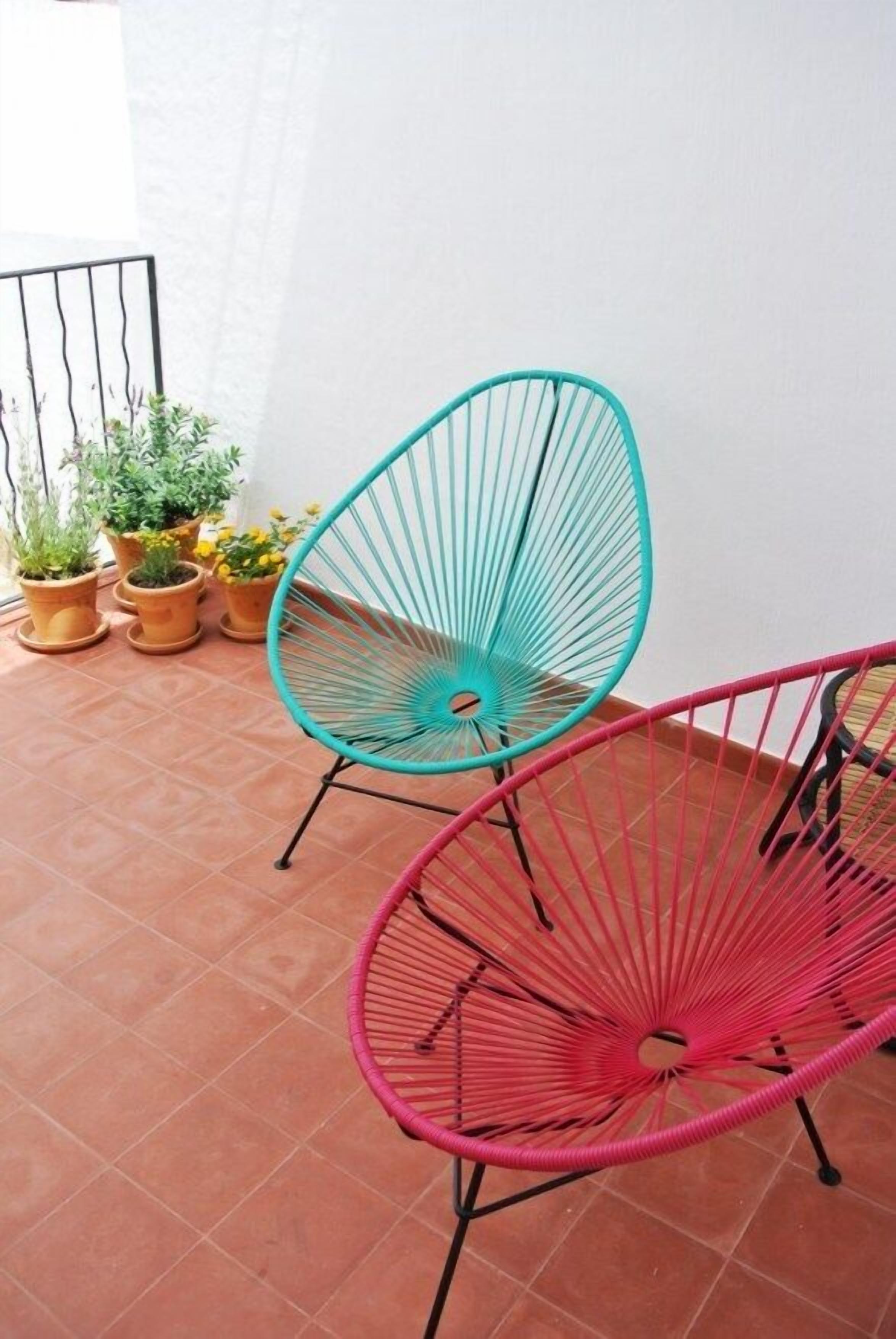 Dos sillas de esquema, una turquesa y otra rosa, sobre un patio con baldosas y plantas en macetas.