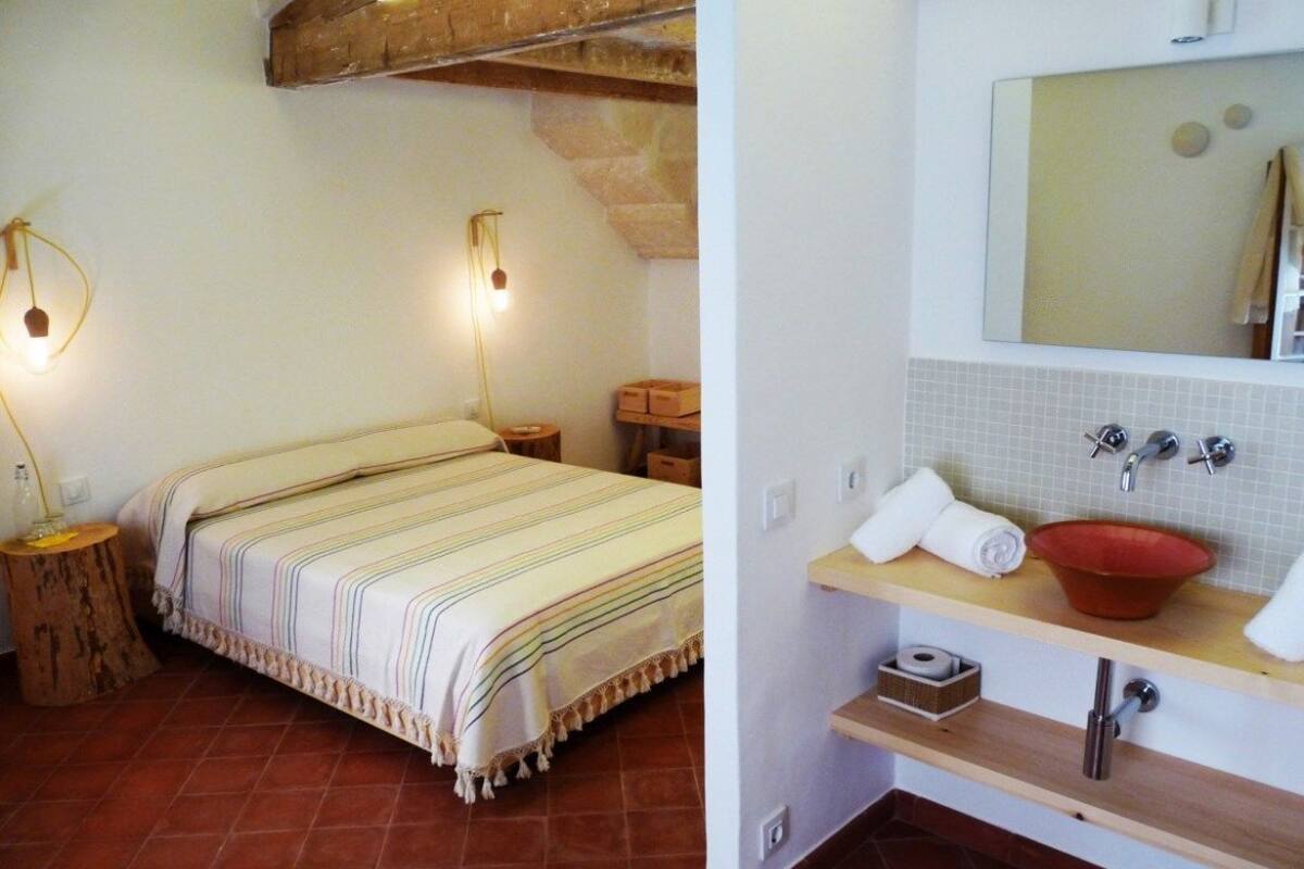 Une chambre à coucher avec un lit, des lampes de chevet et un plafond avec une poutre en bois rustique. La salle de bain est équipée d’un lavabo, d’un miroir et de murs carrelés.