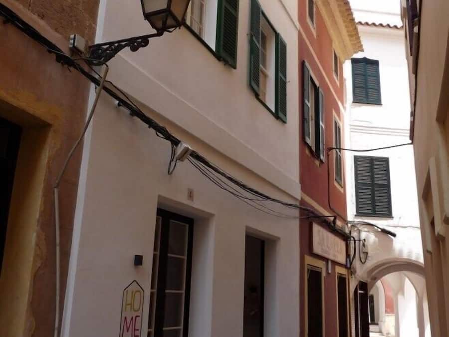 A narrow street with buildings featuring traditional architecture, including shuttered windows and a sign that reads "HO ME".