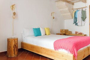Una cama de madera con ropa de cama blanca, un plaid rosa y almohadas coloridas. Hay dos mesitas de noche con lámparas, y un banco de madera con una toalla y un objeto pequeño encima. Ropa cuelga de un perchero sobre la cama.