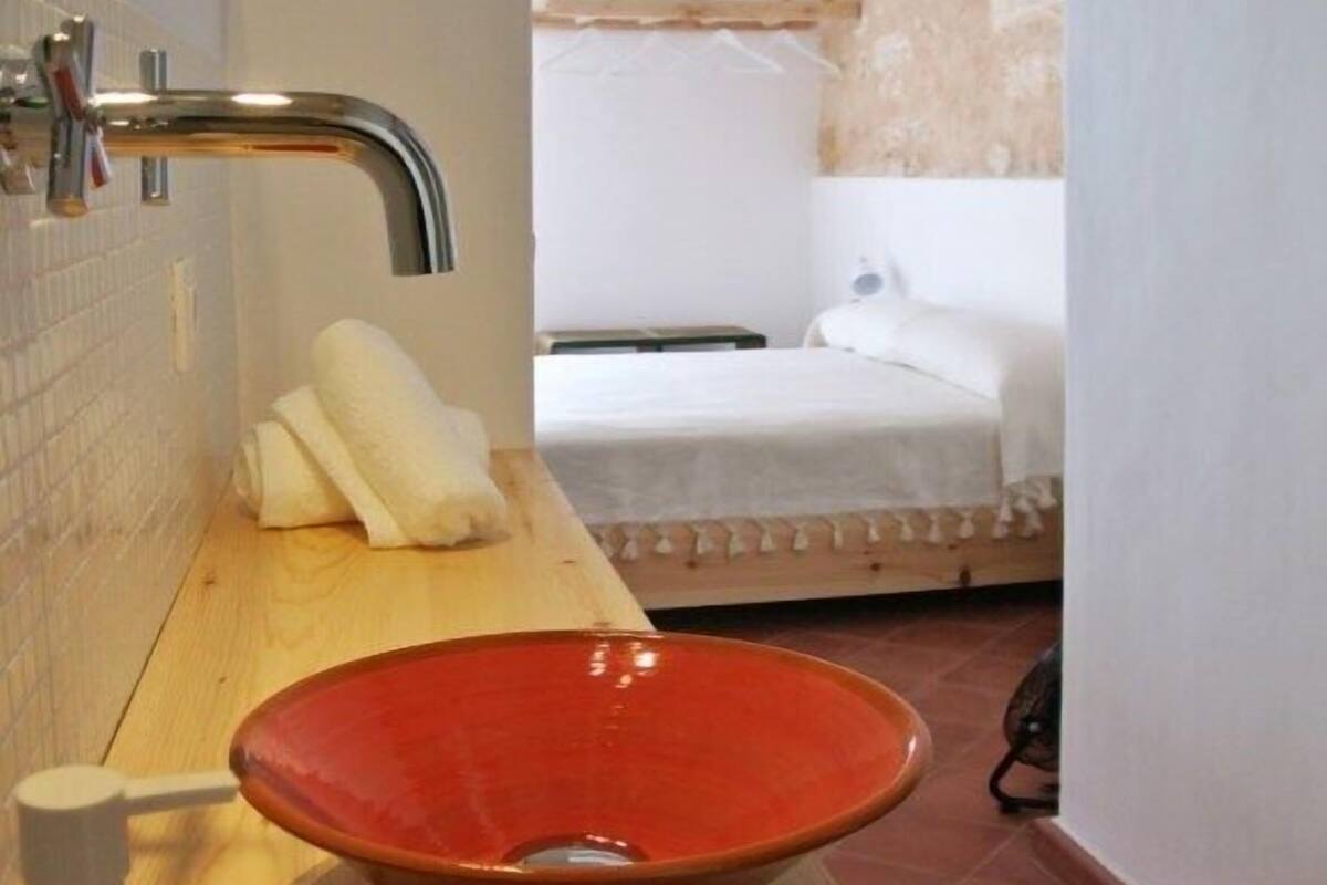Une salle de bain avec un plan de toilette en bois, un lavabo rouge, une serviette blanche et un lit en arrière-plan.