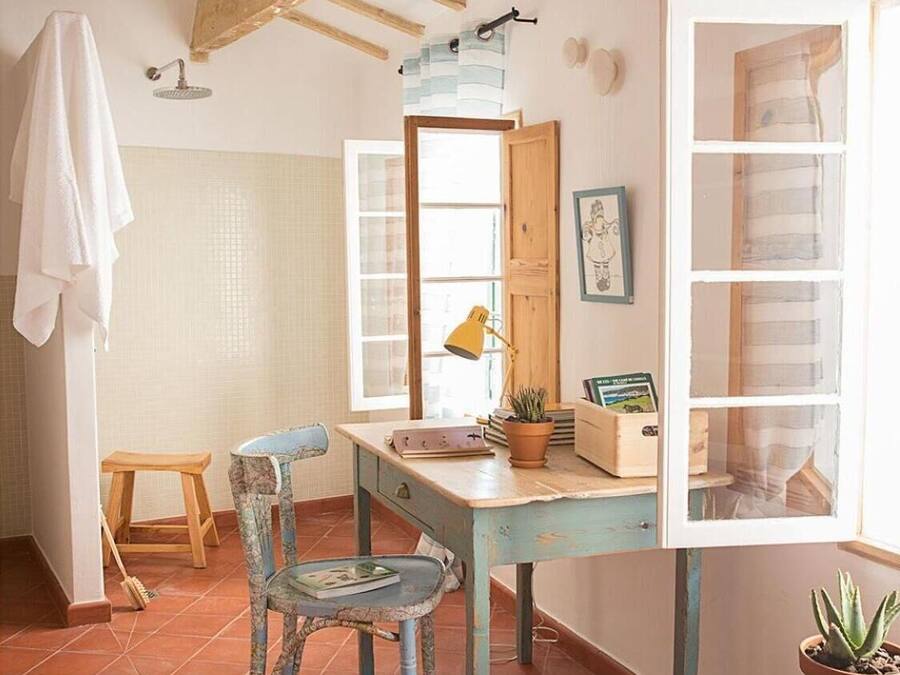 Una habitación rústica con vigas de madera a la vista, suelo de baldosas, un escritorio de madera con una silla, un pequeño taburete, una planta en maceta y una ventana con vistas.