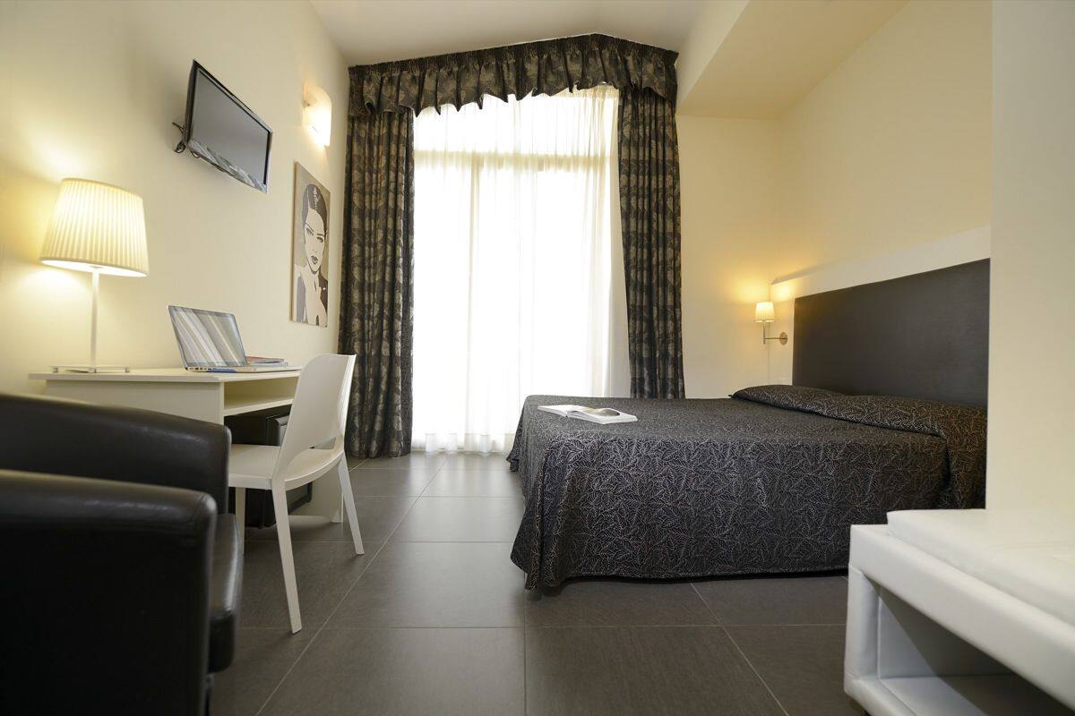 A hotel room with a bed, a desk with a chair, a TV mounted on the wall, and a window with curtains.