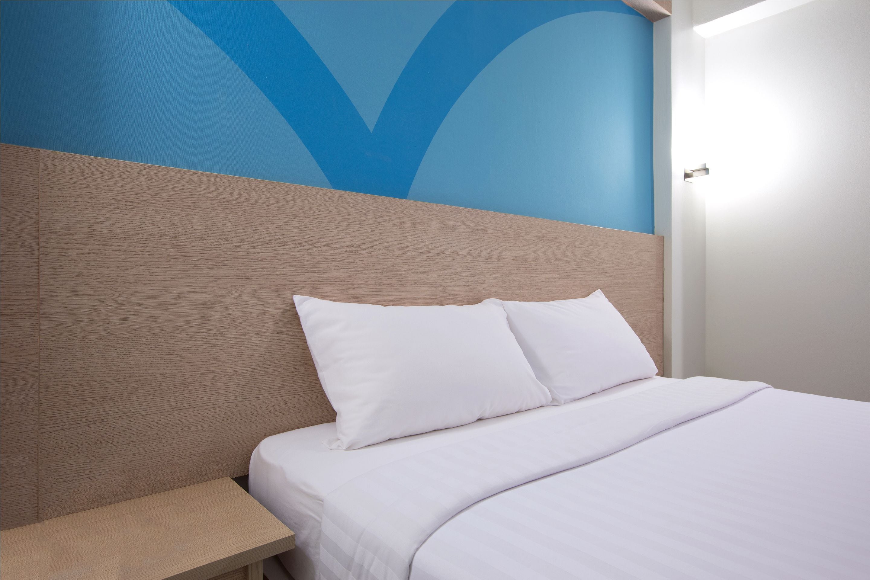 Una cama bien hecha con sábanas blancas y dos almohadas, una mesita de noche de madera a su lado, y una pared con un diseño abstracto en azul.