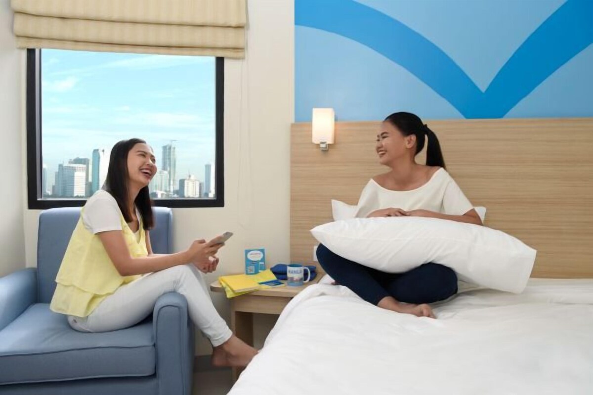 A hotel room with a city view, a bed with white bedding, a nightstand with a lamp, and a window with a view of tall buildings.