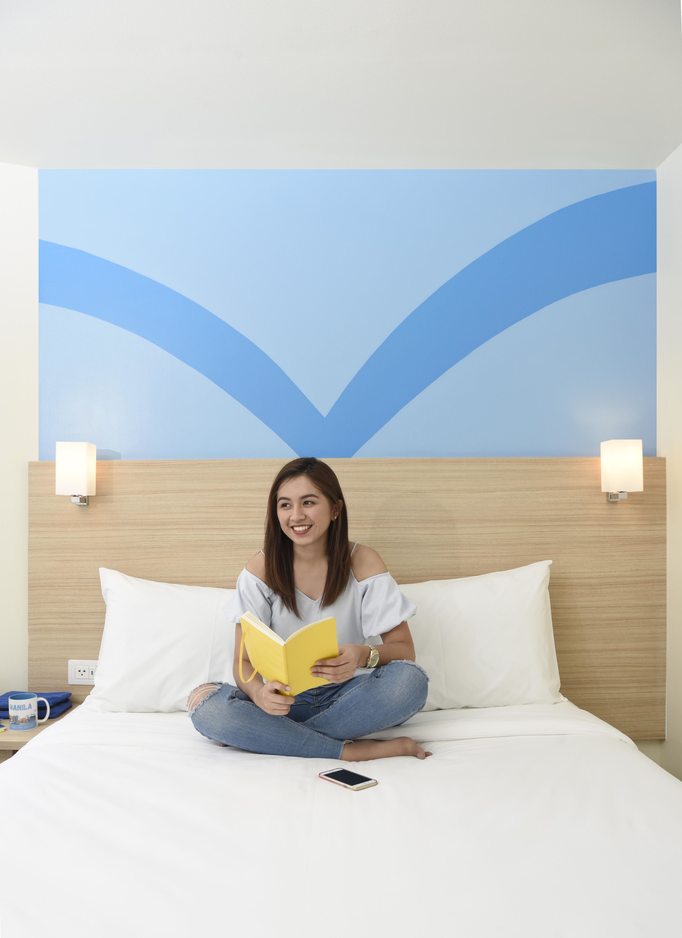 A person sitting on a bed with a book, a phone, and a cup on a bedside table.