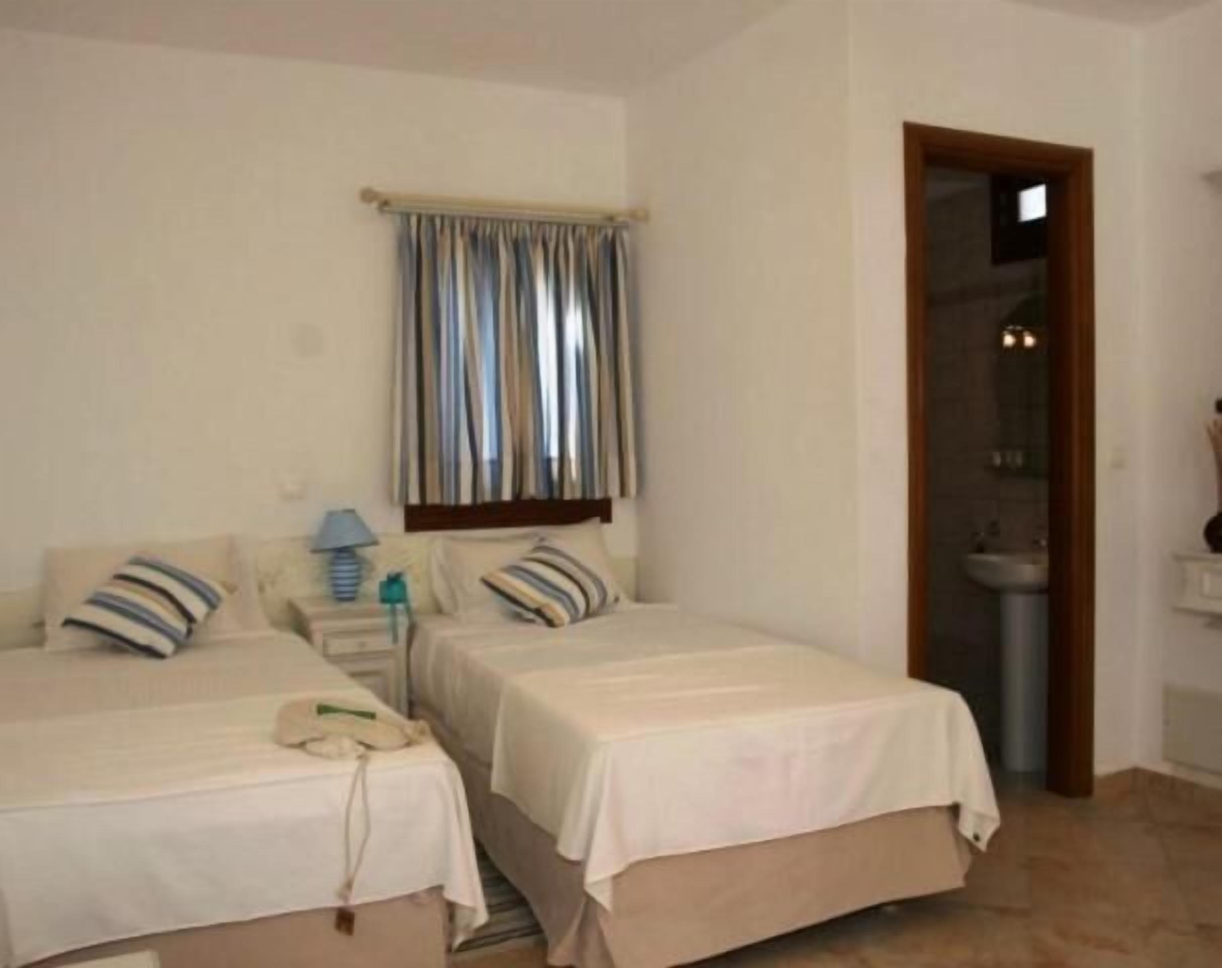 Une chambre d’hôtel avec deux lits, une petite table avec une lampe, un téléphone et une salle de bain visible à travers une porte ouverte.