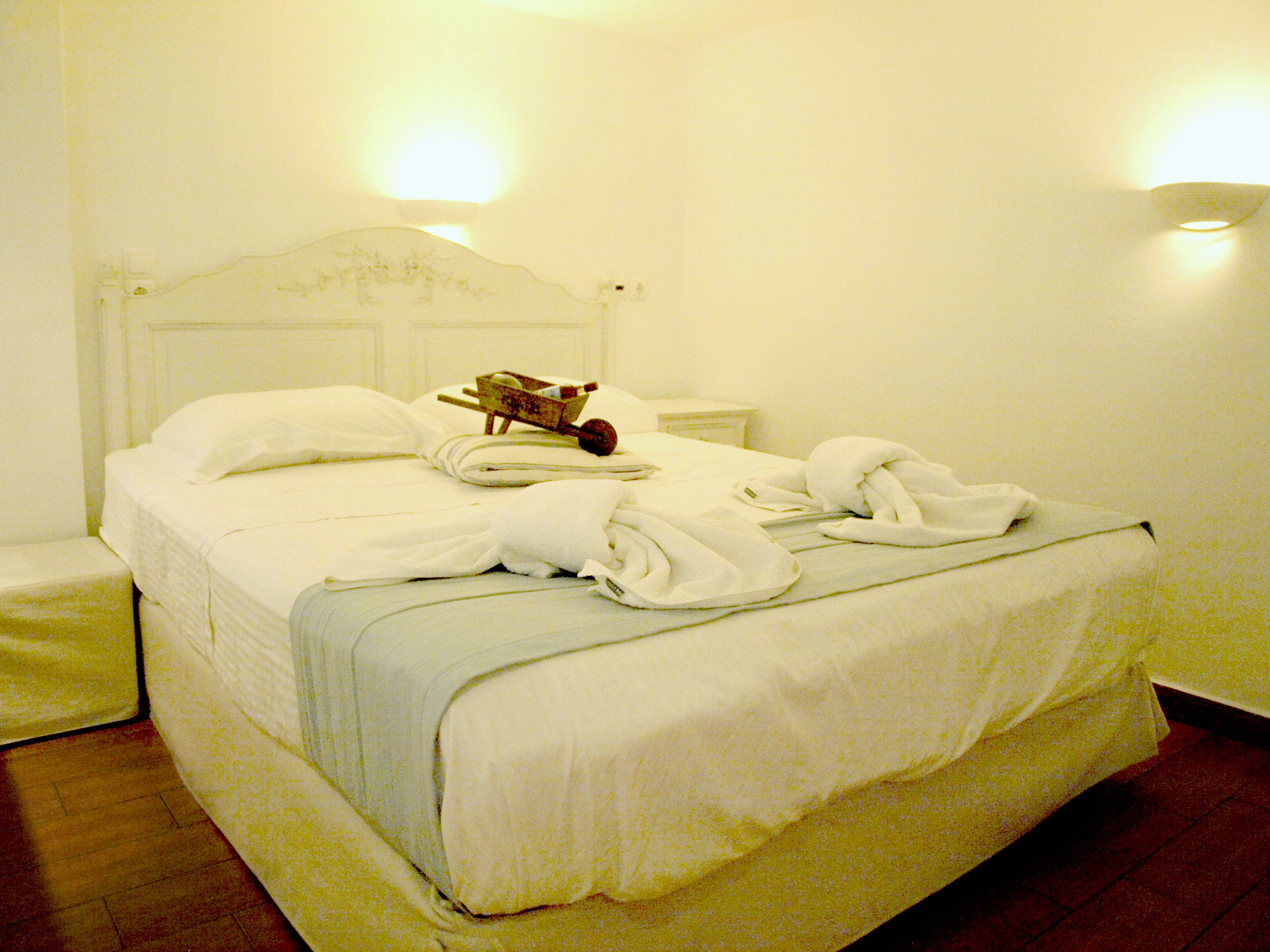 Una cama bien tendida con sábanas y toallas blancas, una cesta de madera y una mesita de noche con una lámpara.